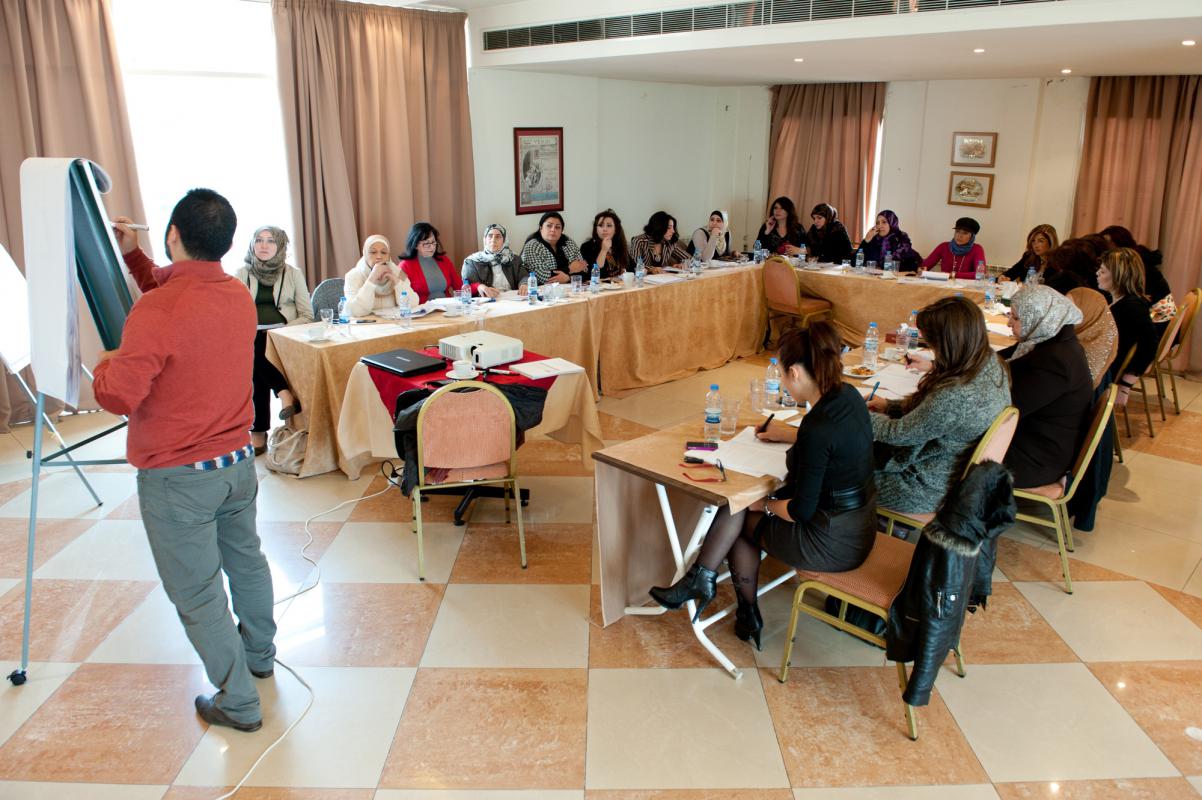 A group of professionally dressed women sit around a table while a man presents to them using an easel