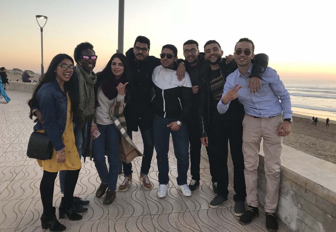 A large group of students poses on the boardwalk at the beach. The sun is setting over the ocean behind them.