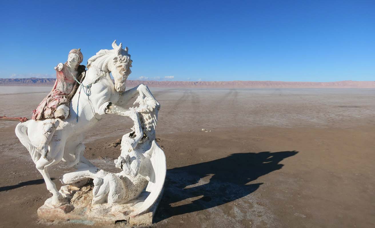 Ruins of an old statue of a person on a rearing horse sits in the foreground with empty plains stretching out behind and a blue sky.