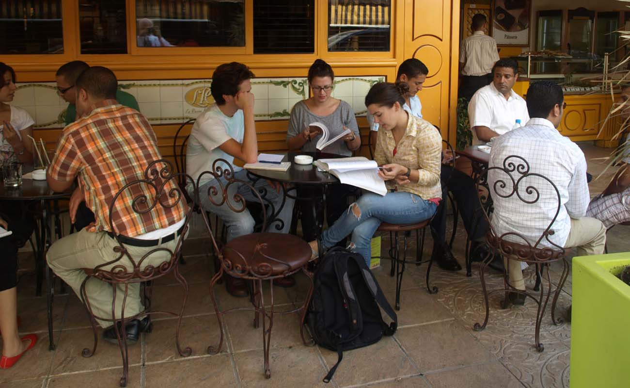 A group of three students studies at a table in front of a busy cafe. The cafe has orange walls