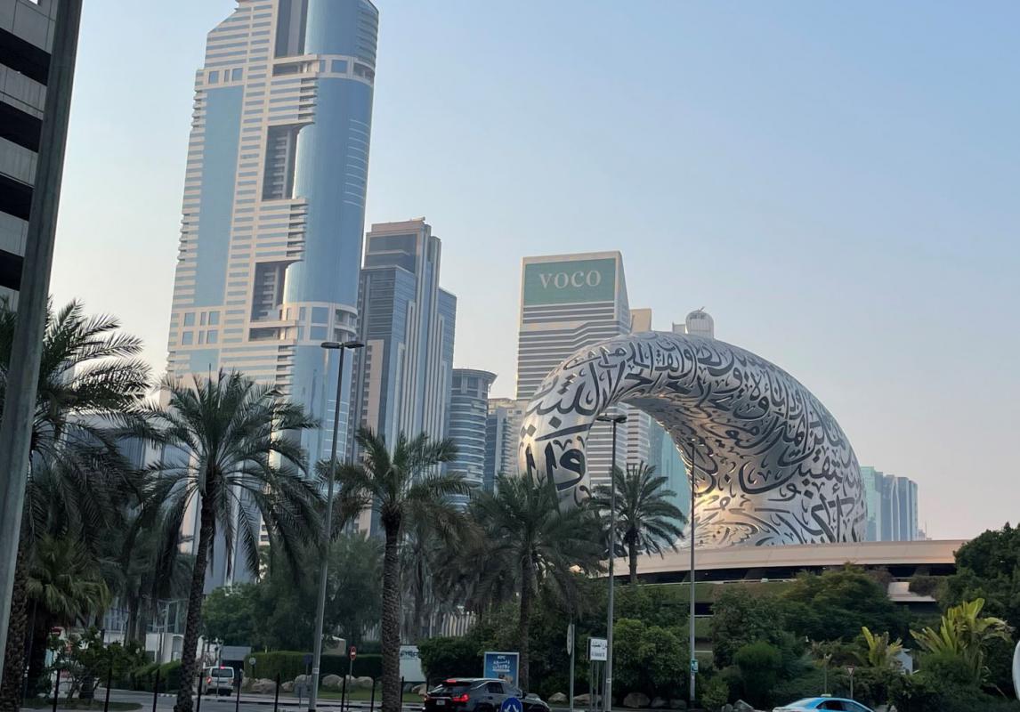 This view from Dubai shows a blue sky day with several skyscrapers in the background. The building on the left is the tallest and they get smaller from there. The focal point of the photo is a large metal arch that is covered in black Arabic calligraphy. There are palm trees in the foreground.