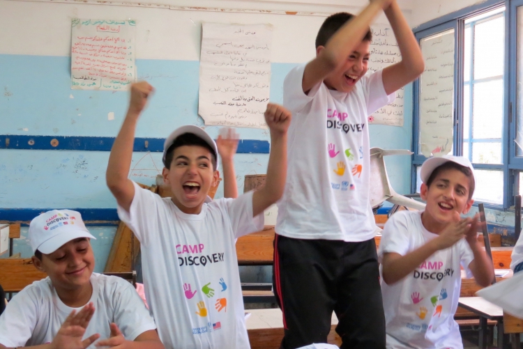 Students wearing Camp Discovery shirts jump and celebrate in a classroom