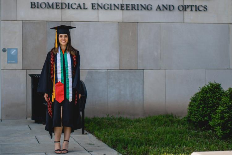 Young woman in graduation robes in front of biomedical engineering and optics building.