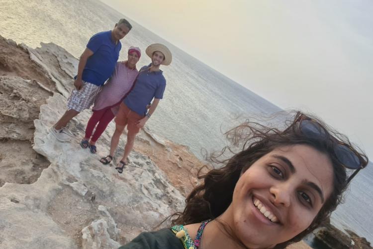 A young woman takes a selfie on the beach. In the background are a middle-aged couple and a young man standing together and smiling for the camera.