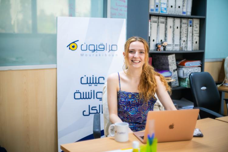 A female student sits at a table behind a Mac Book. She wears a blue tank top and has her copper curly hair in a ponytail, she smiles at the camera. She sits in a library or office, with a bookshelf behind her and a floor stand banner in Arabic for some organization