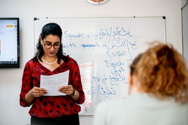 A teacher holds a piece of paper at the front of the class in front of a board with Arabic and English written on it.