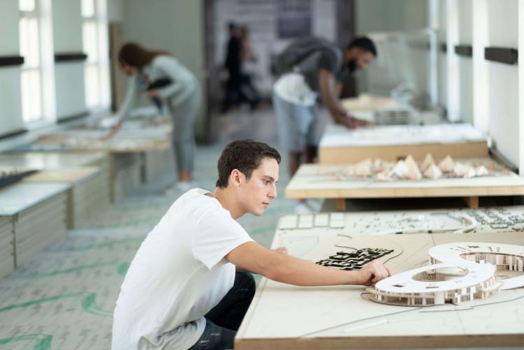 A student crouches on the ground to work on an architectural model placed on a low table. There are several other similar tables in the background.