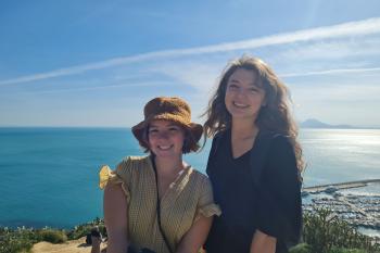 Two women smile from a vantage point that overlooks the blue sea under a sunny blue sky.