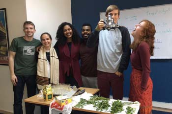 6 students pose in their Arabic classroom during a tea activity. One student is holding up the teapot and the girl beside him is looking up at the teapot. 