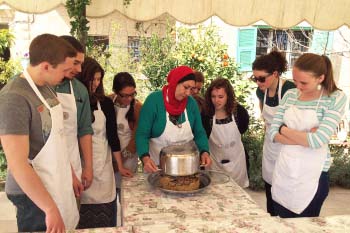 A group of students in aprons stands around a Jordanian woman as she teaches them how to cook. She is flipping over a pot and talking about the process
