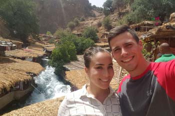 A man and a woman pose together in front of a nature scene with a river. There are some trees and a mountain rising in the background