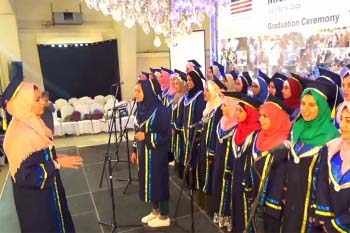 A group of female high school graduates wear their caps and gowns onstage while singing for an audience, one student is using a microphone