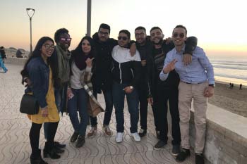 A large group of students poses on the boardwalk at the beach. The sun is setting over the ocean behind them.