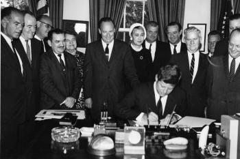 President Kennedy signing a bill with people watching behind him