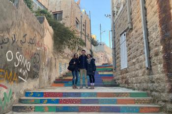 Three students pose on a colorful staircase between buildings in Amman. They are in the middle of the photo on a landing. The wall on the left side has graffiti on it.