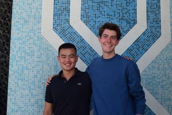 Two men smile at the camera while standing in front of a mosaic tiled wall.