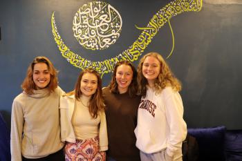 Four women stand together and smile at the camera in front of a dark colored wall with a calligraphy mural on it. 