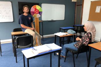 A student sits on a desk in the front of the class presenting to his teacher who is sitting at a student desk in front of him. They are both smiling.