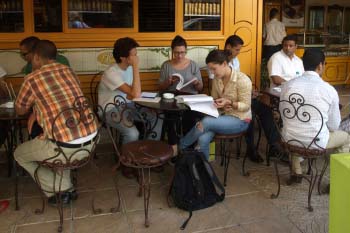 A group of three students studies at a table in front of a busy cafe. The cafe has orange walls
