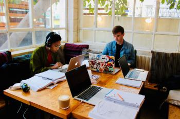 Two students study at a table in a cafe. The table is covered with papers and laptops