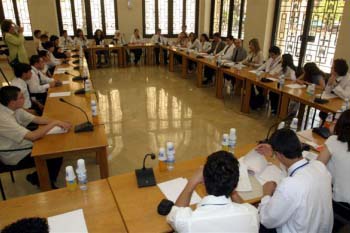 A large group of students sit at multiple long tables facing each other with paper and microphones