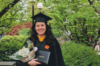 A graduating student wears a cap and gown and holds a diploma and flowers