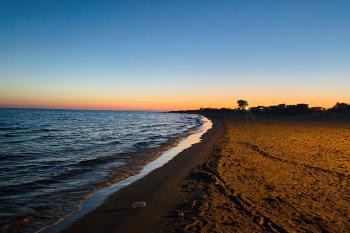 The sun sets over the beach that is on the left of the frame and the sand is on the right and there are no people