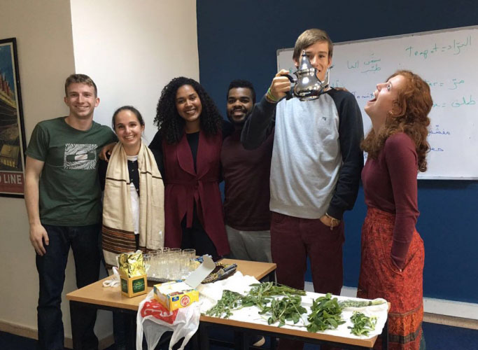 6 students pose in their Arabic classroom during a tea activity. One student is holding up the teapot and the girl beside him is looking up at the teapot. 