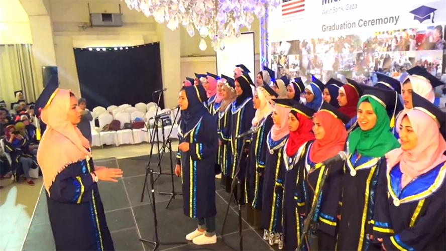 A group of female high school graduates wear their caps and gowns onstage while singing for an audience, one student is using a microphone