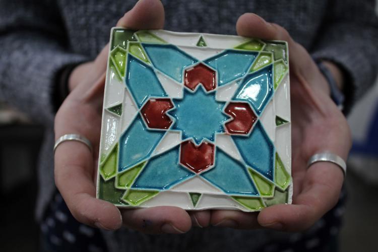 A student holds a square mosaic tile that they made