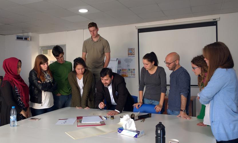 Students stand around their calligraphy teacher who is showing them how to write