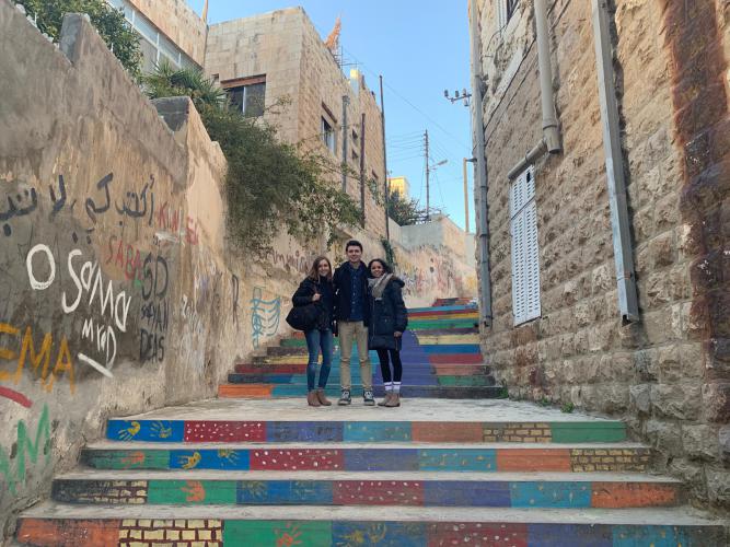 Three students pose on a colorful staircase between buildings in Amman. They are in the middle of the photo on a landing. The wall on the left side has graffiti on it.