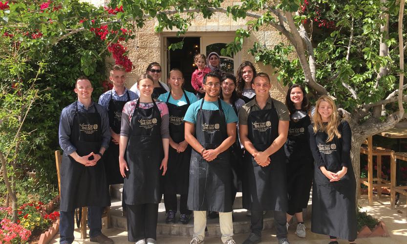 A group of students in black branded aprons smiles at the camera in front of a Jordanian home and plants.