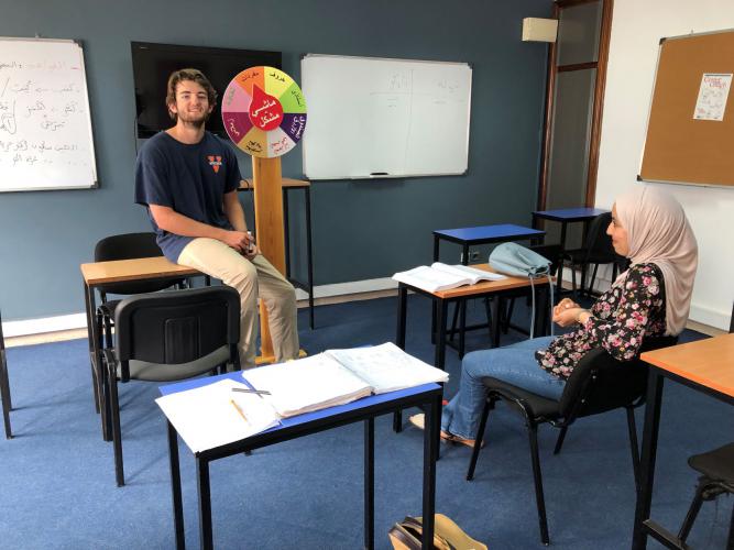 A student sits on a desk in the front of the class presenting to his teacher who is sitting at a student desk in front of him. They are both smiling.