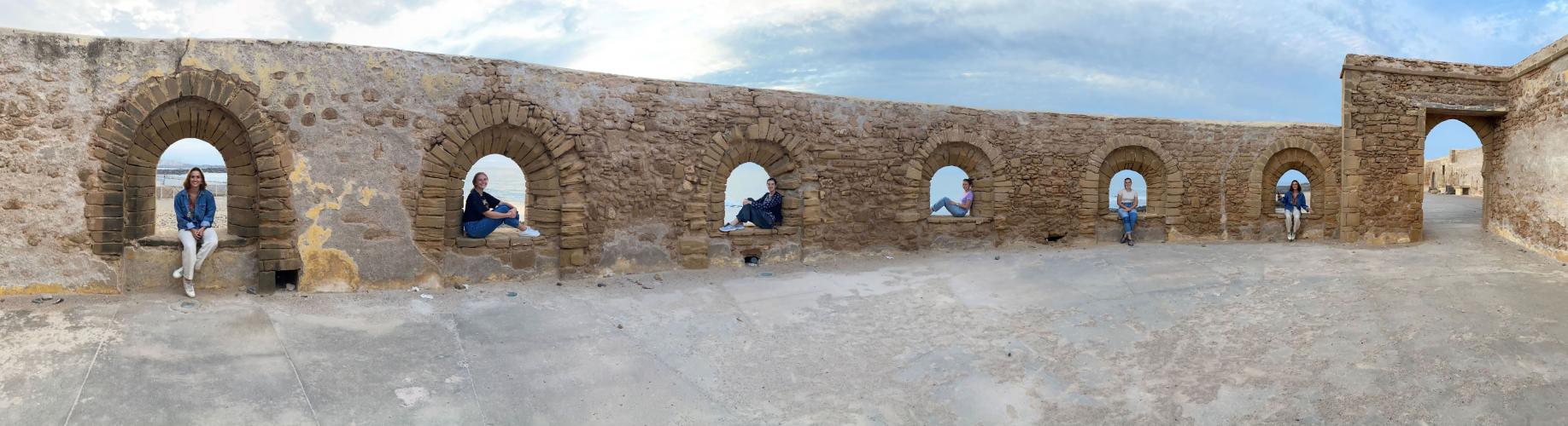 Students sit inside separate arches in a panorama image of an old wall.