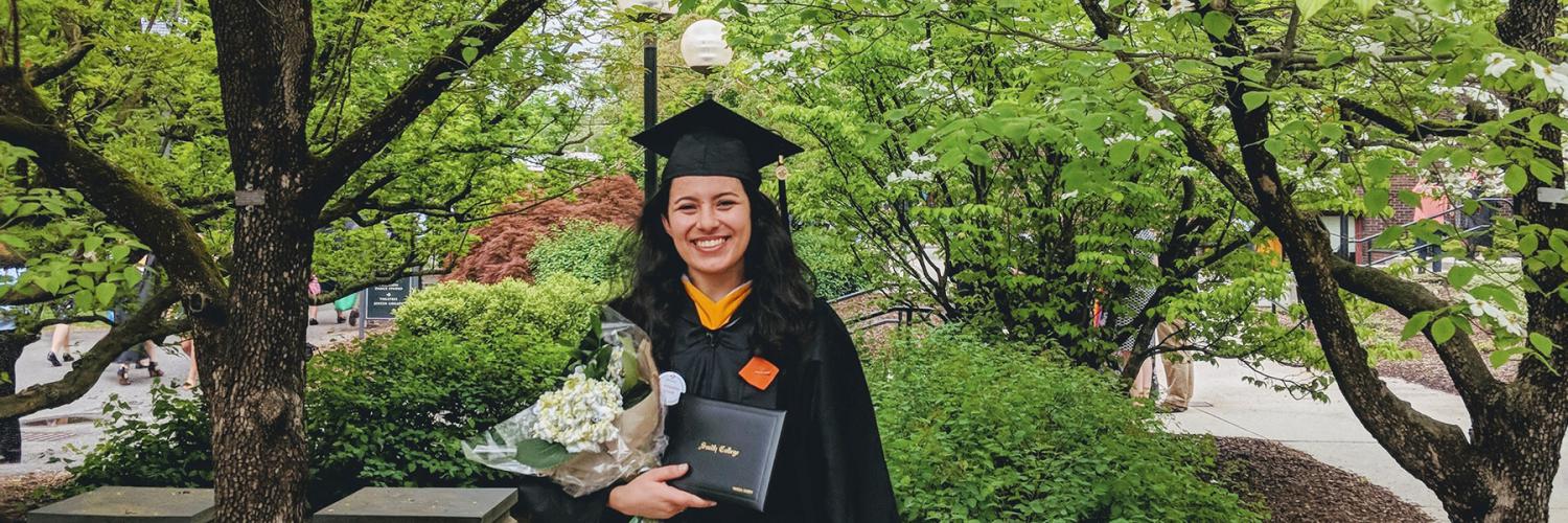 A graduating student wears a cap and gown and holds a diploma and flowers