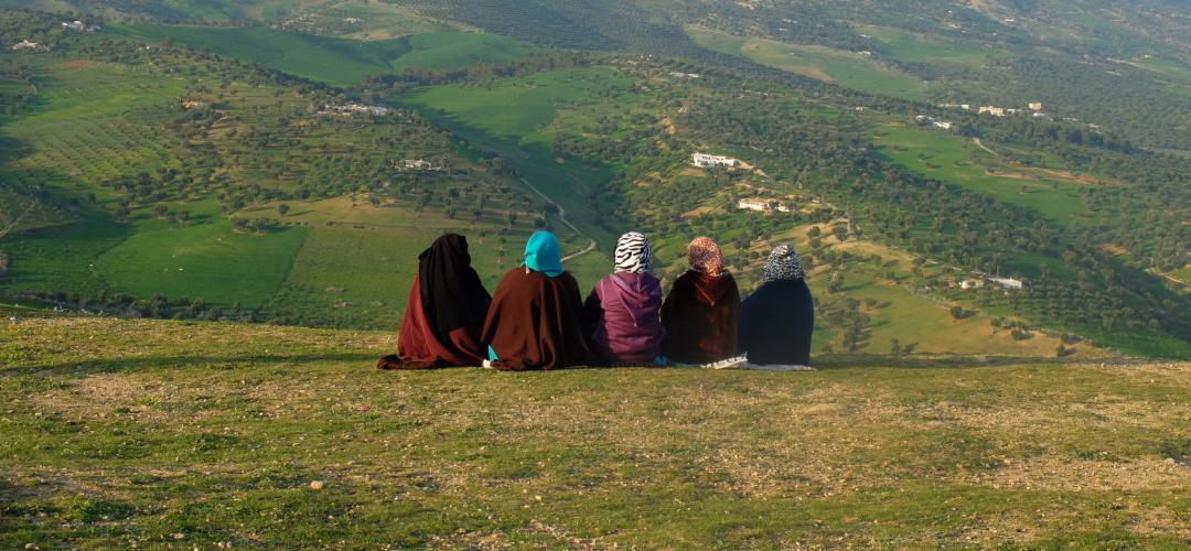 Five women in traditional Moroccan clothing and hijabs sit on the crest of a hill looking out towards the fields and hills beyond.