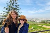 Two women stand a smile in front of a vista of green fields and city in the background.