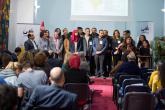 A large group of BLADI participants stand on a stage in front of an audience while one woman speaks into a microphone