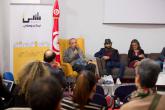 A man sits in a chair onstage while speaking into a microphone, the BLADI logo and a Tunisian flag are behind him