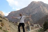 A man stands on a rock and opens his arms wide as he smiles at the camera which is below him. Behind him rises a mountain and between him and the mountain is a small town.