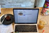 A laptop is open to a podcast site with a cup of iced coffee on one side and a piece of cake on the other. A notebook and pens are scattered around the laptop.