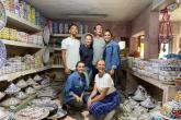 6 people pose for the camera in a pottery shop with finished products all around them on shelves waiting to be sold.