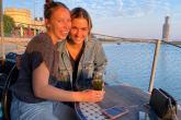 Two women sit together alongside the river drinking tea. The setting sun lights their smiling faces.