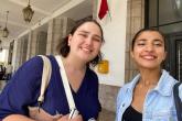 Two women pose together and smile for a selfie. They are both smiling and are standing in front of a large building.