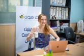 A student holds a coffee cup and smiles in front of a banner for an industry and she is sitting at a table with a laptop