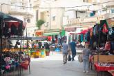 Stalls of clothes and related good line the street between off-white buildings. A few people walk down the street