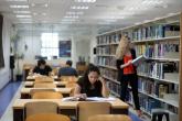 Inside the university library one woman reads a book from off of the shelf while several students sit and study at different tables.