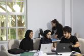 Three students study together. There are two women on the left and a man on the right. They have laptops on the table and are talking.