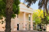 A building on campus with pillars sits in the sun with palm trees in front and around.